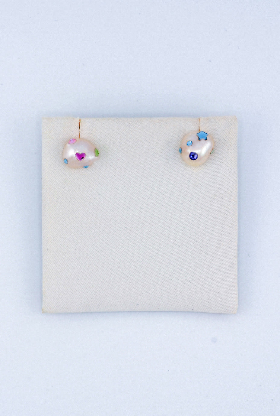 A pair of cute, light-colored pearl handmade earrings displayed on a soft beige backing. One earring is decorated with a pink heart shaped Austrian crystal and a green shaped Austrian crystal, while the other has blue crystals, including a star shaped and a purple Austrian crystal.