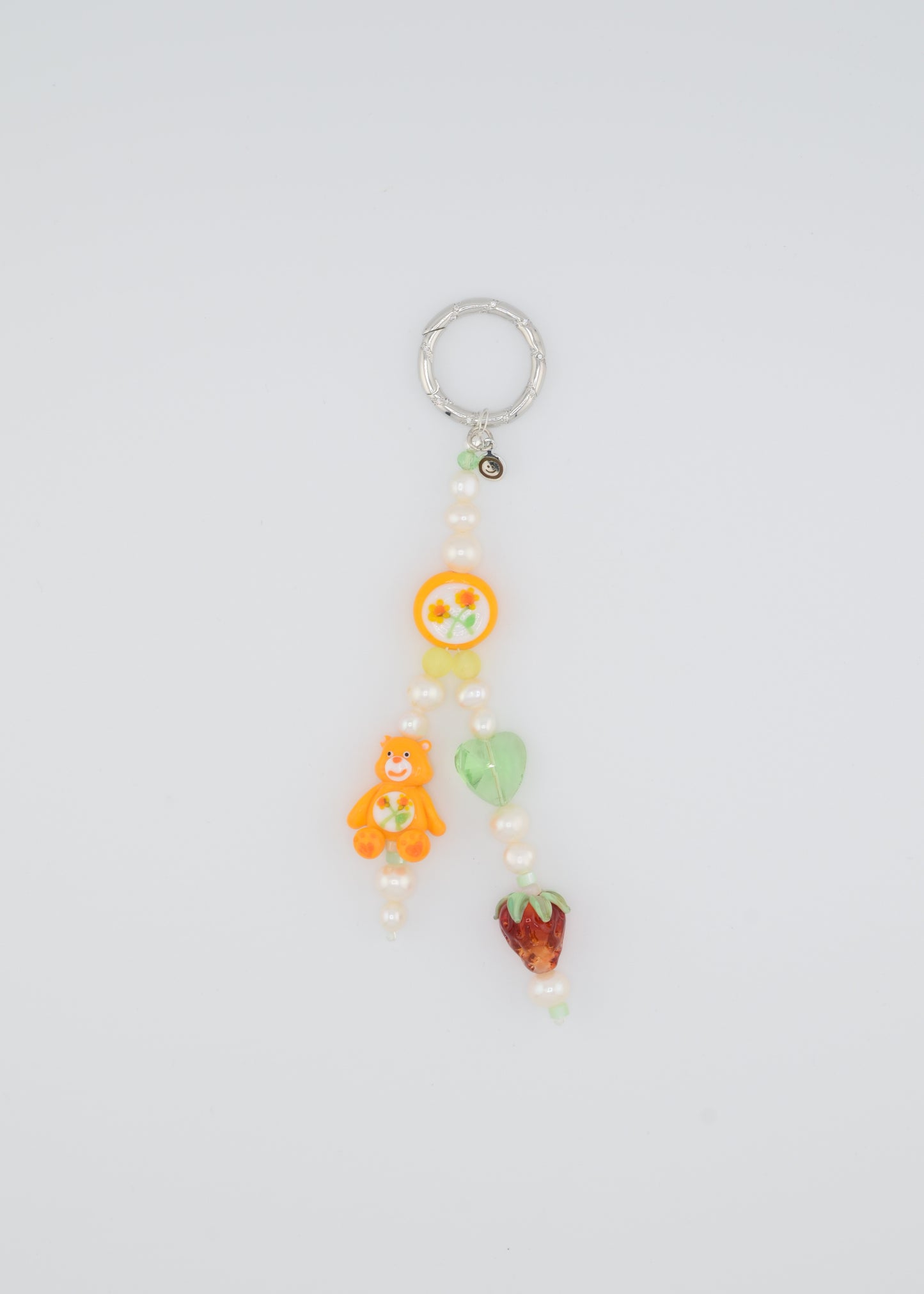A colorful beaded bag charm featuring freshwater pearls, an orange Care Bear lampwork bead, a hand-painted floral bead, a green glass heart bead, and a glass strawberry bead. The charm is attached to a silver keyring and has a playful, nostalgic aesthetic.