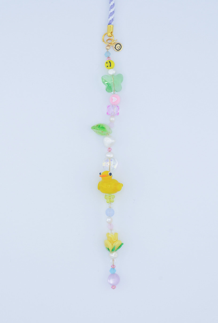 A close-up shot of the phone strap featuring a cheerful smiley face bead, a butterfly, flower, and leaf glass lampwork bead, along with a cute yellow duck bead, a green glass heart bead, and vibrant pastel-colored beads. A playful and retro for phone accessories.