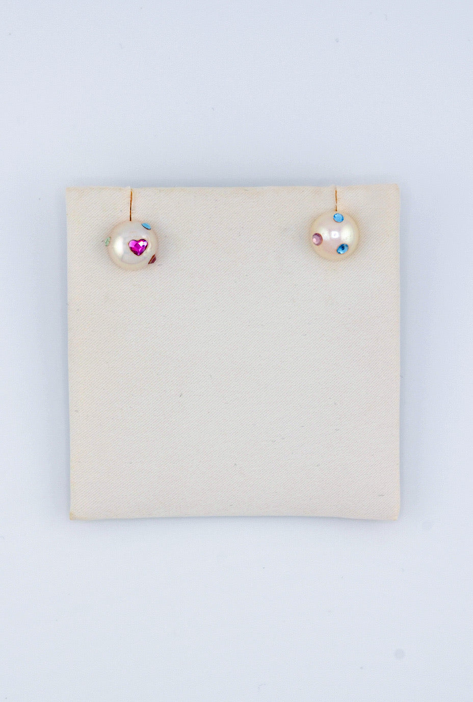 These playful, handmade pearl earrings are decorated with colorful Austrian crystal gems. One earring features a shiny pink heart and a green gem, while the other is adorned with a cluster of blue and purple crystal gemstones, creating a whimsical design.