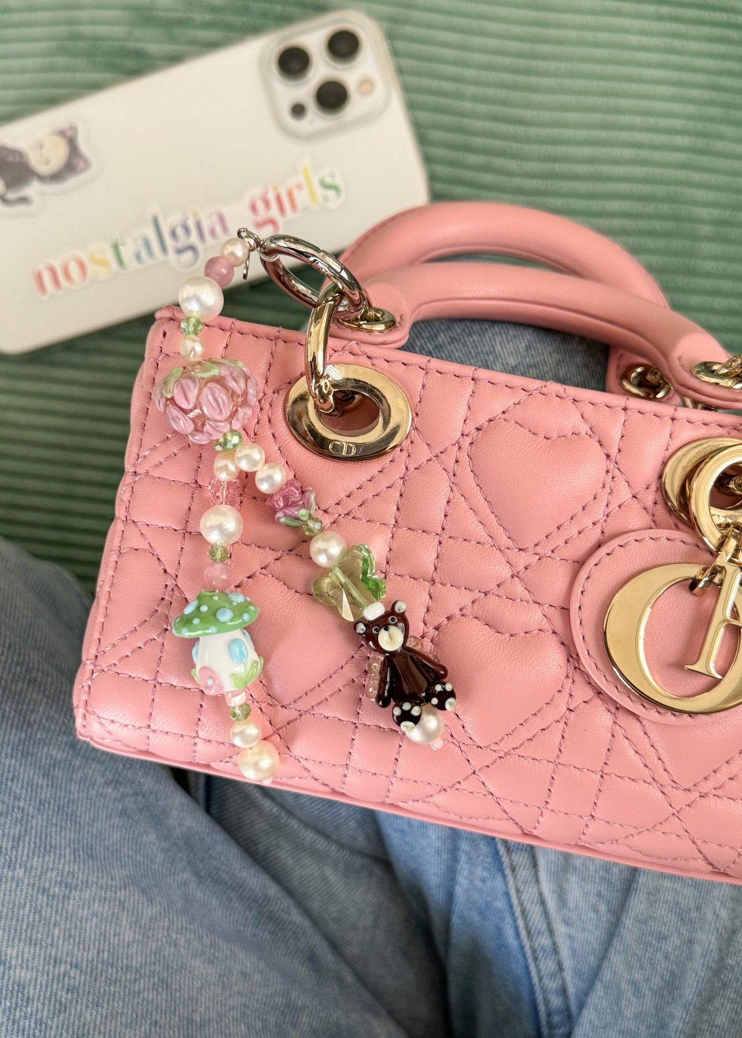 The bag charm is attached to the handle of a pink quilted handbag with gold hardware. The charm dangles from one of the bag’s gold rings, adding a whimsical touch to the structured purse. In the background, a smartphone with a pastel-colored "nostalgia girls" case is resting on a green fabric surface. The setting gives a cozy and stylish feel.
