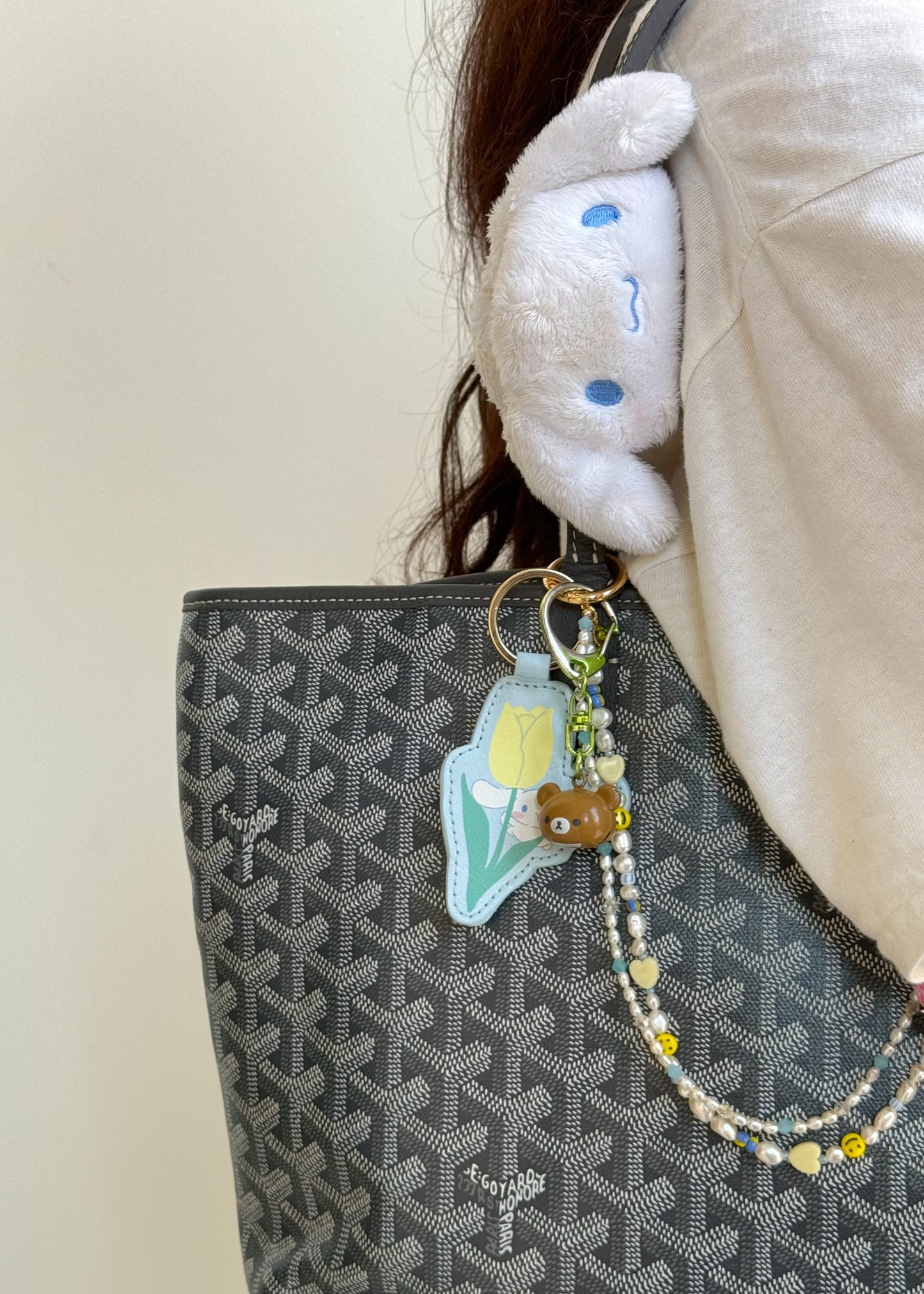A bag with a patterned black and gray design, featuring the beaded bag charm hanging from a gold clasp. The charm is attached to a keyring that also holds a green metal clip, a tulip-shaped leather tag, and a small brown bear figurine. A white Sanrio character claw clip with blue eyes is hooked onto the bag’s strap.