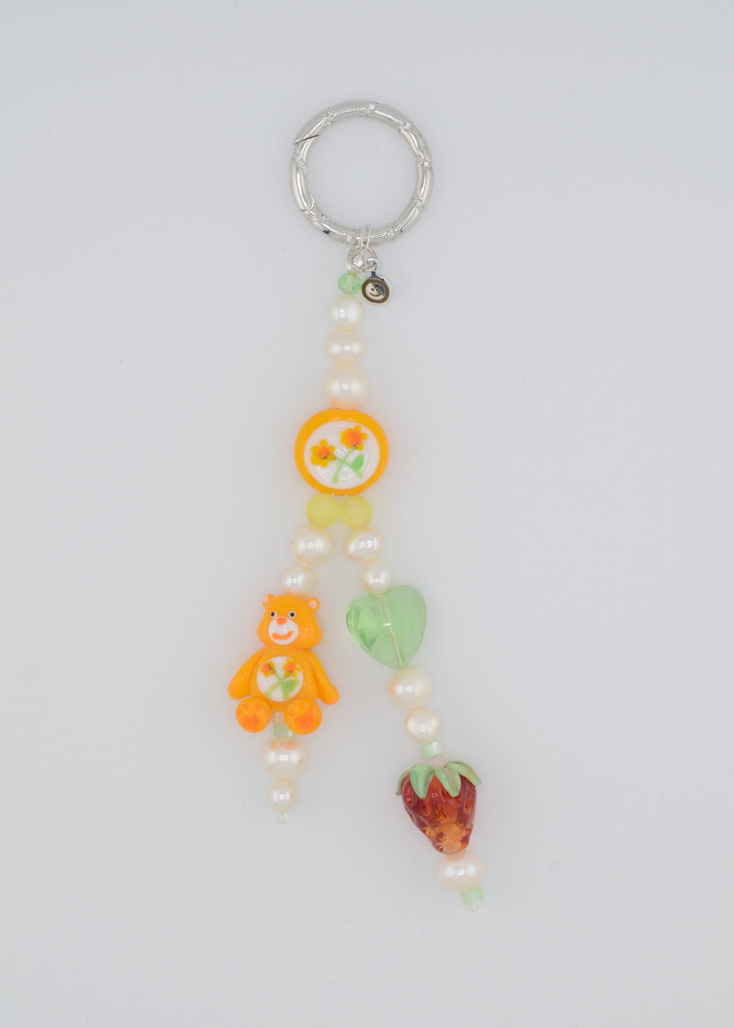 A colorful beaded bag charm featuring freshwater pearls, an orange Care Bear lampwork bead, a hand-painted floral bead, a green glass heart bead, and a glass strawberry bead. The charm is attached to a silver keyring and has a playful, nostalgic aesthetic.