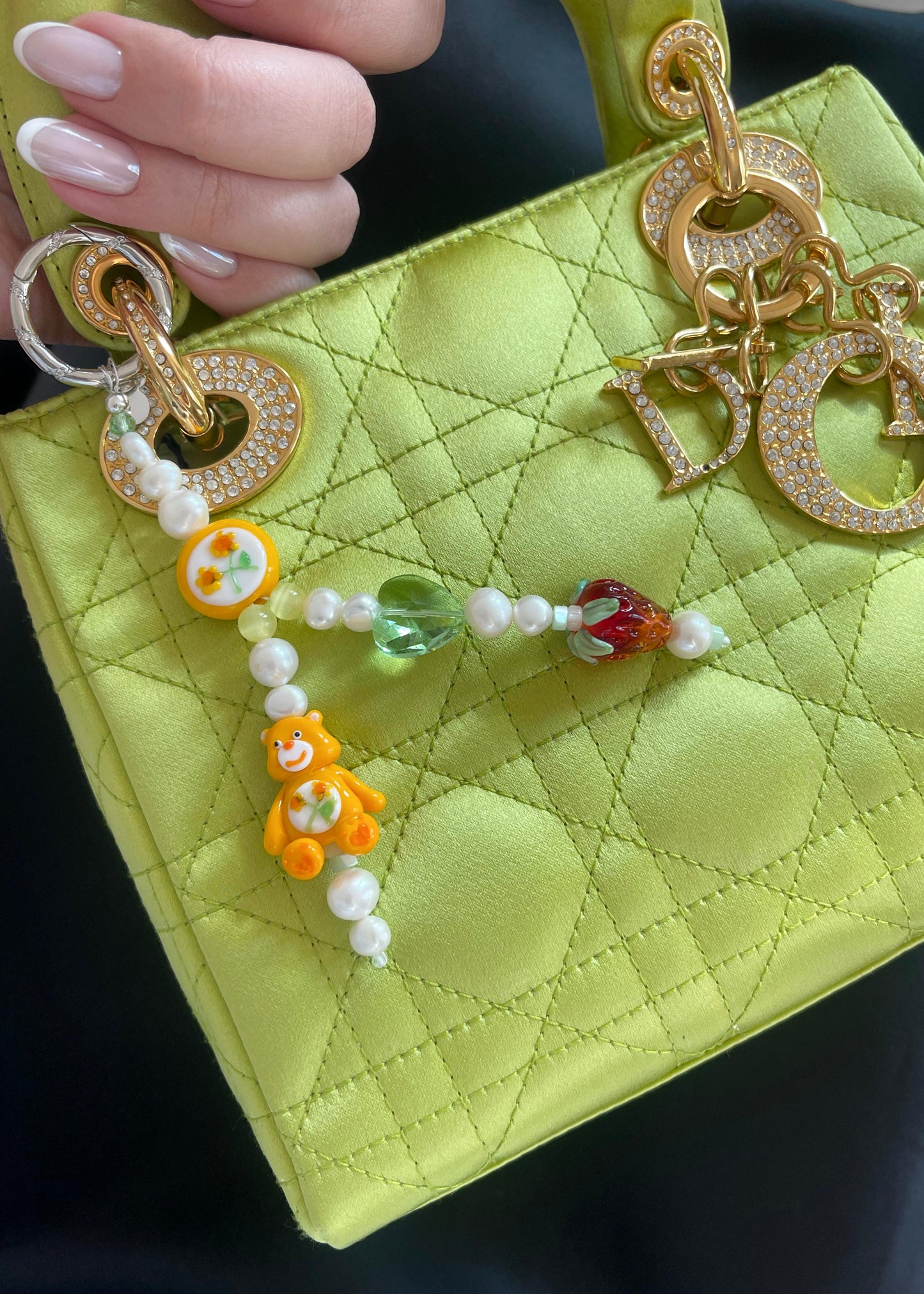 A close-up of a lime green quilted handbag with gold hardware and a rhinestone-studded "Dior" charm. Attached to the bag is a beaded bag charm featuring freshwater pearls, an orange Care Bear lampwork bead, a hand-painted floral bead, a green glass heart bead, and a glass strawberry bead. A hand with a French manicure holds the bag, complementing its elegant and playful aesthetic.