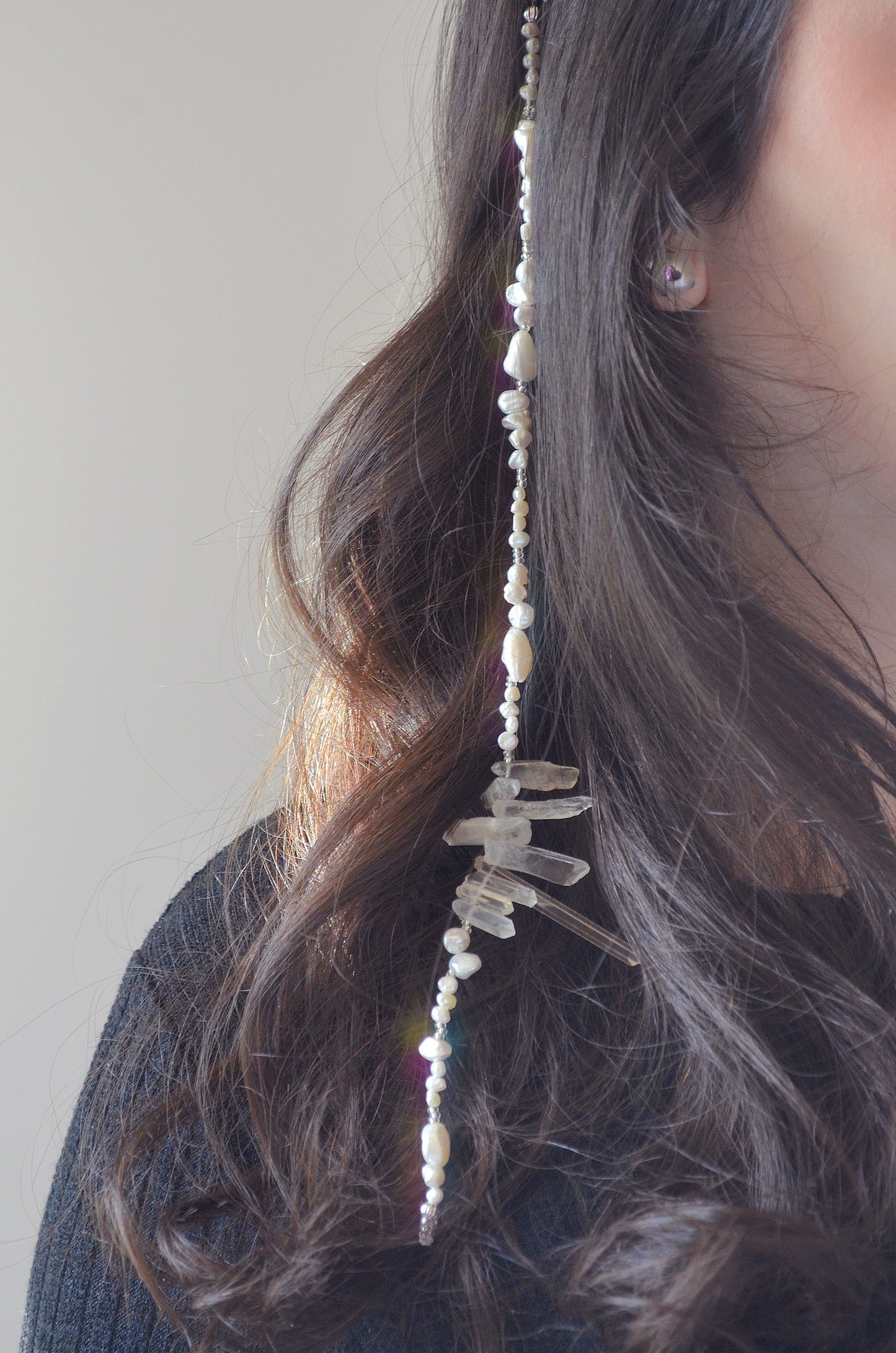 A photo of the hair accessory worn in a female's dark, wavy hair. The accessory, made of freshwater pearls and clear quartz, is positioned along a section of hair, with the quartz crystals clustered in the center and pearls trailing down. The light catches the pearls, making them appear iridescent