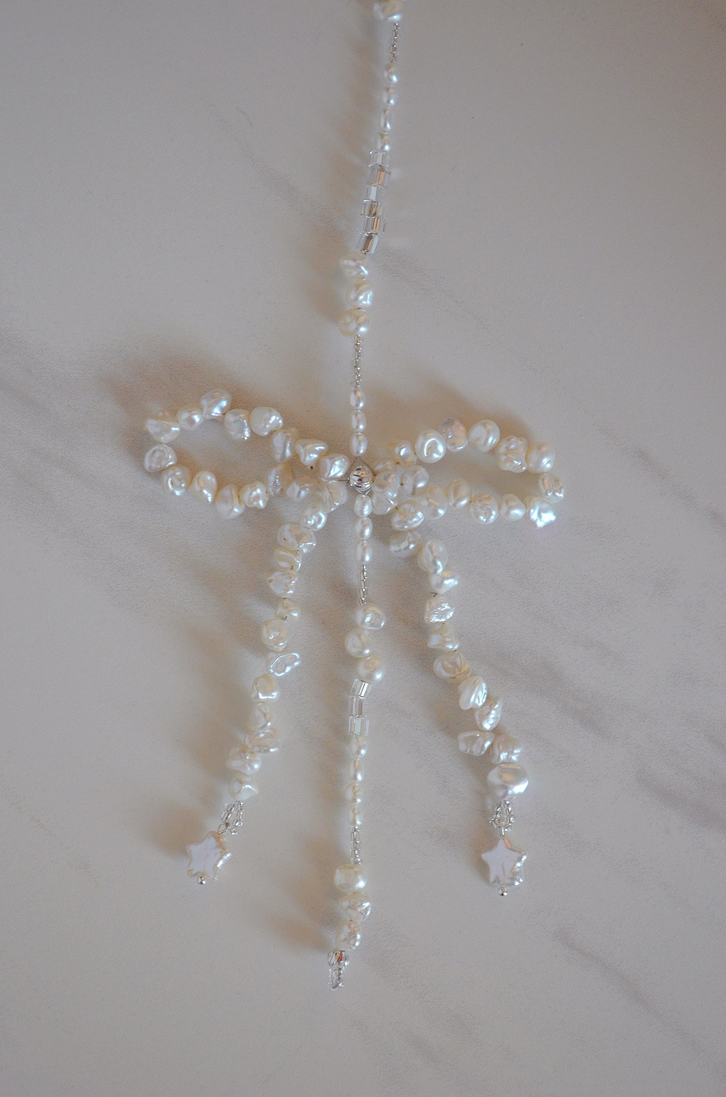 A close up shot of the bow at the bottom of the hair accessory made up of genuine freshwater pearls and silver beads against a white and grey marble background. The bottom of the bow features star shaped pearls.