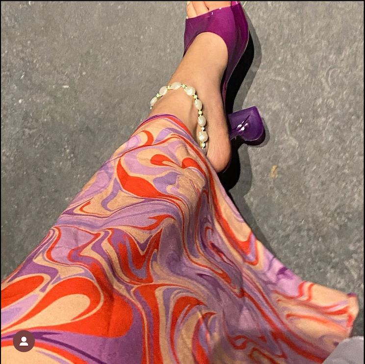 A stylish female adorned with the white seashell anklet, accented with vibrant turquoise beads, paired perfectly with a flowy purple and orange patterned dress and purple heels for a bold, boho chic look.