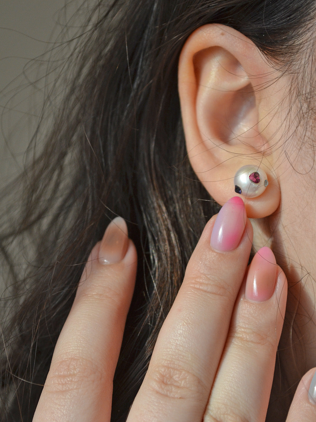 Whimsical pearl earrings with bright accents, showcasing a pink heart and blue Austrian crystal gemstones, worn by the model as they adjust the earring.