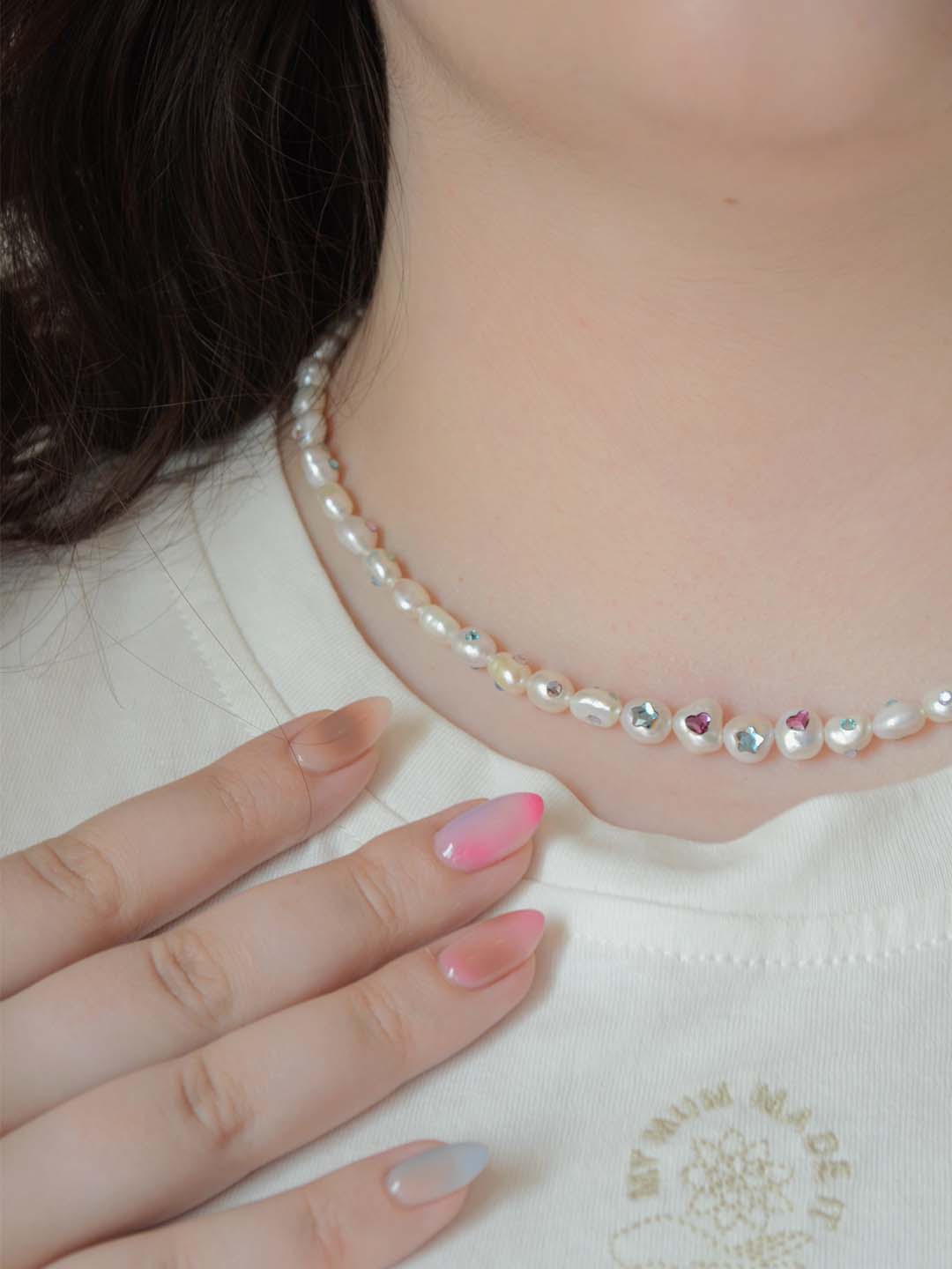 A pearl necklace with colorful Austrian crystal gemstone embellishments worn on a model’s neck. The necklace features playful details such as blue stars, pink hearts, and a gold-tone clasp. The model also wears a light-colored t-shirt.