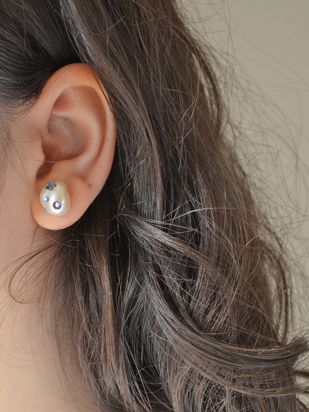 A close-up shot of a woman wearing one of the pearl earrings from the first image. The earring is decorated with blue Austrian crystals, including a star and a purple gem, and is visible on her ear with her hair styled loosely.