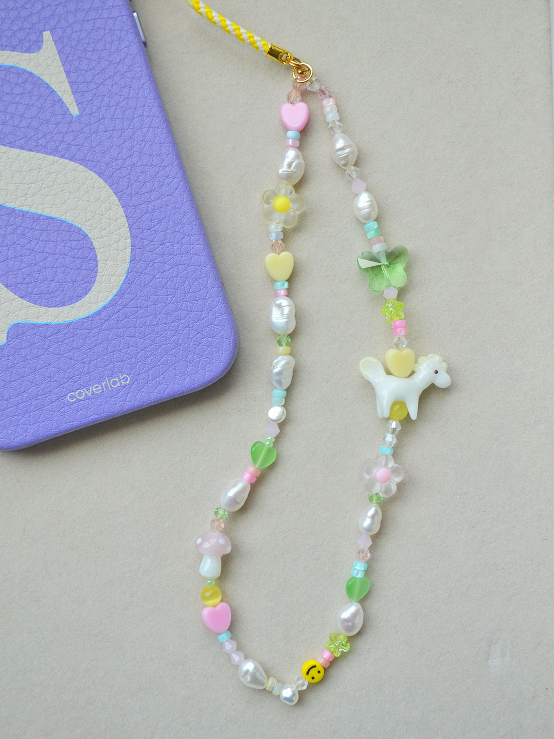 The phone charm is displayed next to a lavender phone case, emphasizing the cheerful and bright design. The charm includes colorful glass beads, freshwater pearls, and a cute pony lampwork bead, making it a playful and eye-catching accessory for your phone.