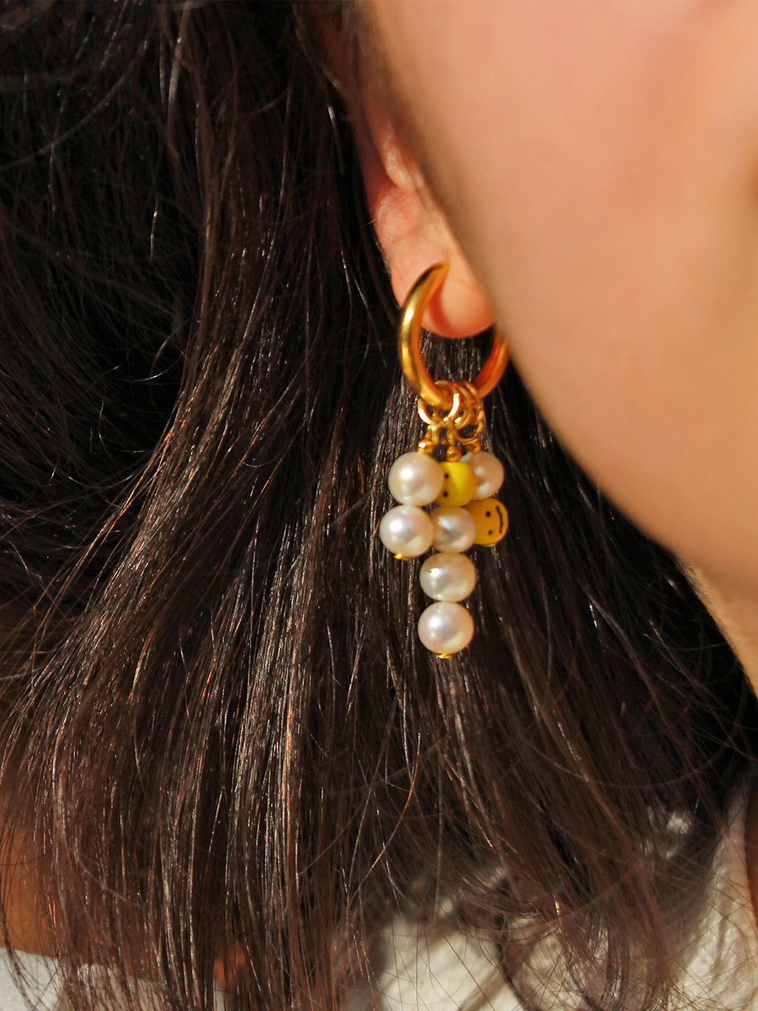 A model shows off a pair of hoop earrings, featuring a mix of pearls and playful yellow smiley face beads, adding a touch of joyful energy to the look.