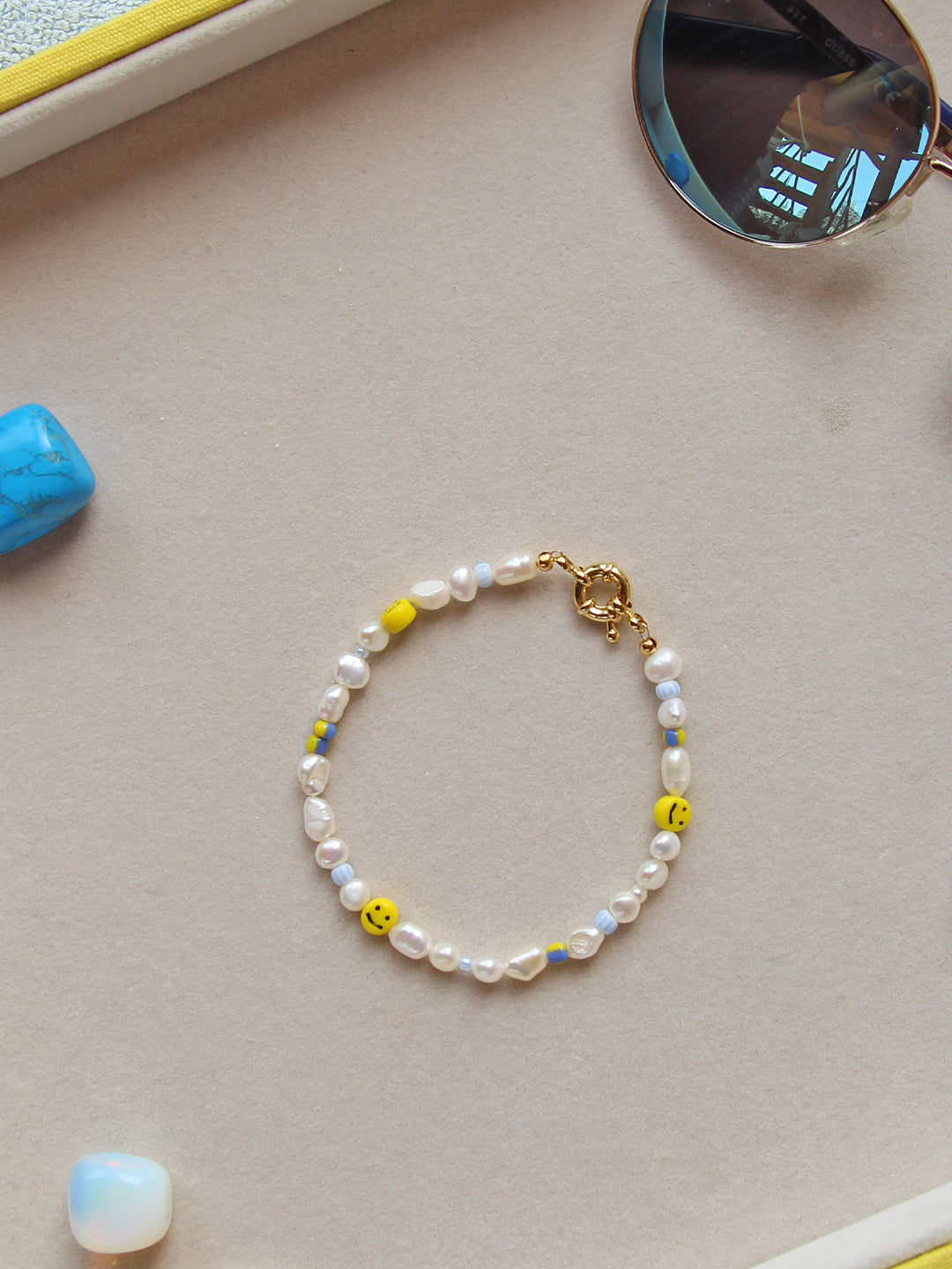 A playful, smiley face bracelet paired with opalescent stones, a few colorful beads, and freshwater pearls. The bracelet has a delicate gold clasp and is styled with sunglasses and other accessories.